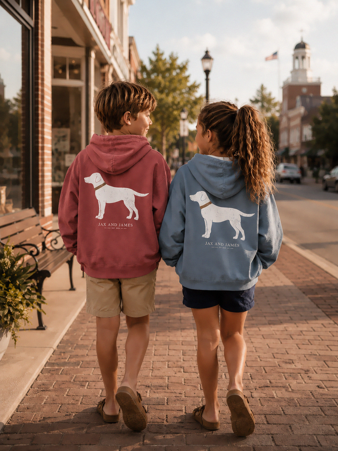 Two children walking hand in hand down a brick sidewalk in front of a storefront, wearing matching hoodies with white dog silhouettes. The child on the left wears a blue hoodie, and the child on the right wears a light pink hoodie.