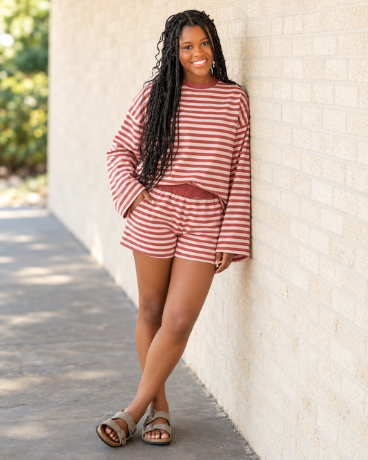 A woman with long black braids stands against a beige brick wall wearing a matching long-sleeve top and shorts set with horizontal rust-red and white stripes, paired with brown sandals.