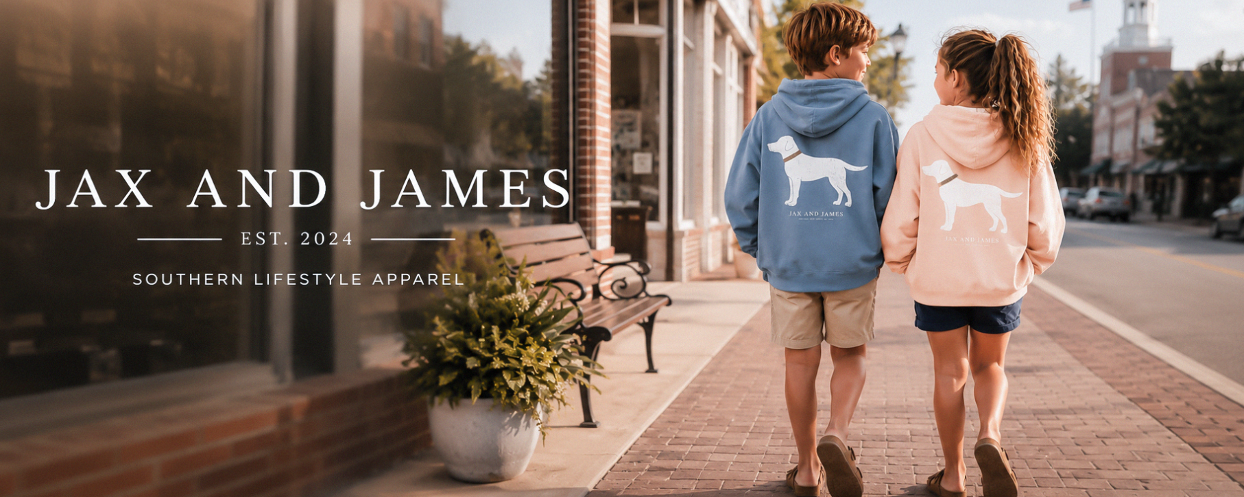 Two children walking hand in hand down a brick sidewalk in front of a storefront, wearing matching hoodies with white dog silhouettes. The child on the left wears a blue hoodie, and the child on the right wears a light pink hoodie.